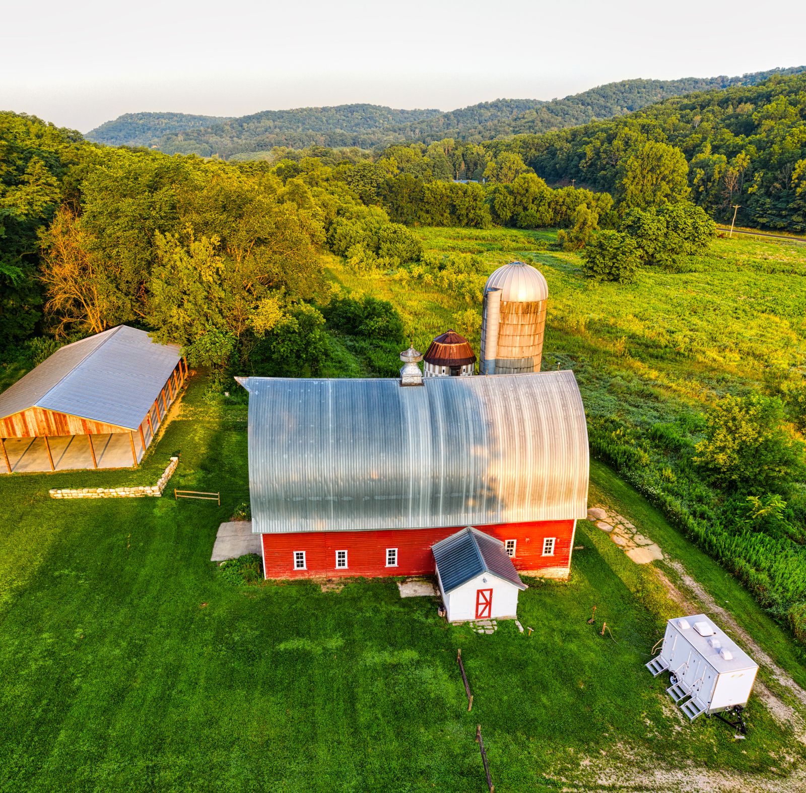 Elevated drone view of rustic barn and open-air pavilion grounds surrounded by green fields and wooded hills
